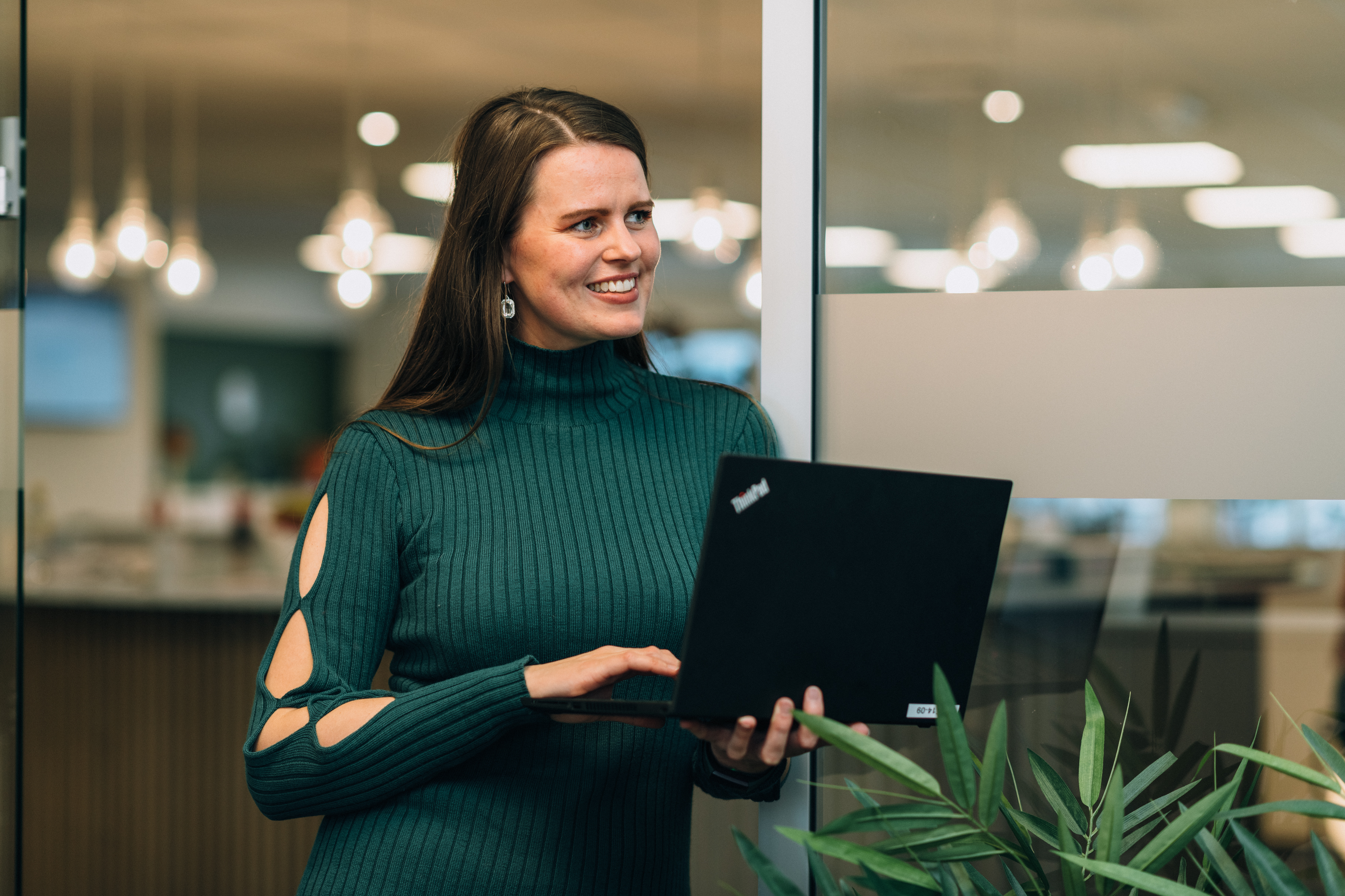 Kvinne i grønn kjole holder en bærbar PC innendørs, med uskarpe lys og planter i bakgrunnen.