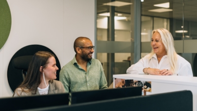 Three people talking in an office, two seated at desks with monitors, one standing and leaning on a partition, with glass walls and office lighting in the background.