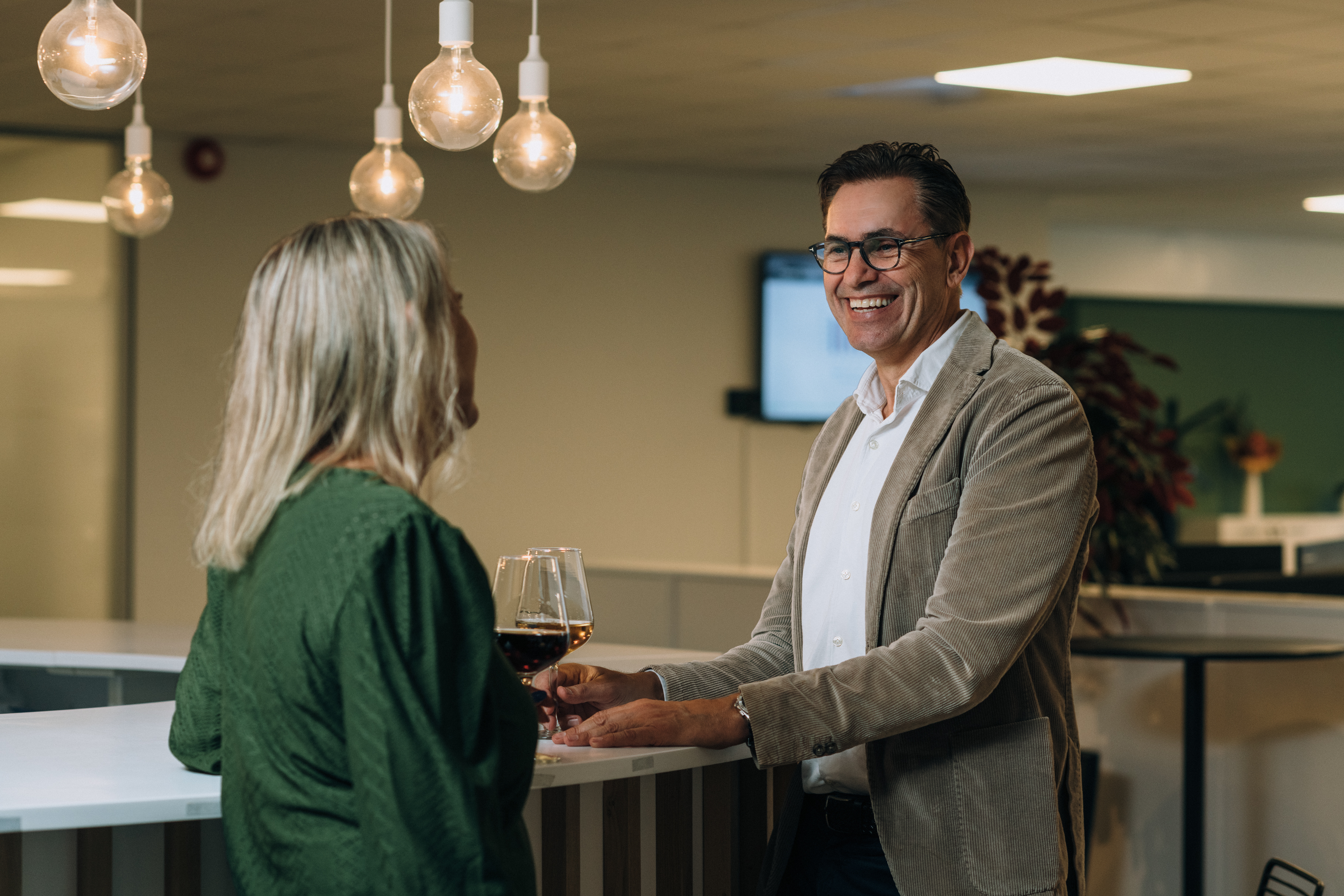 To personer står ved en bar og samtaler innendørs, begge holder hvert sitt vinglass. Hengende lyspærer, en uskarp skjerm og planter er i bakgrunnen.