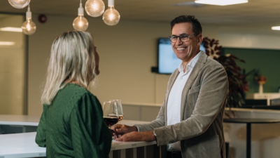 Two people stand by a bar and converse indoors, each holding a wine glass. Hanging light bulbs, a blurred screen, and plants are in the background.