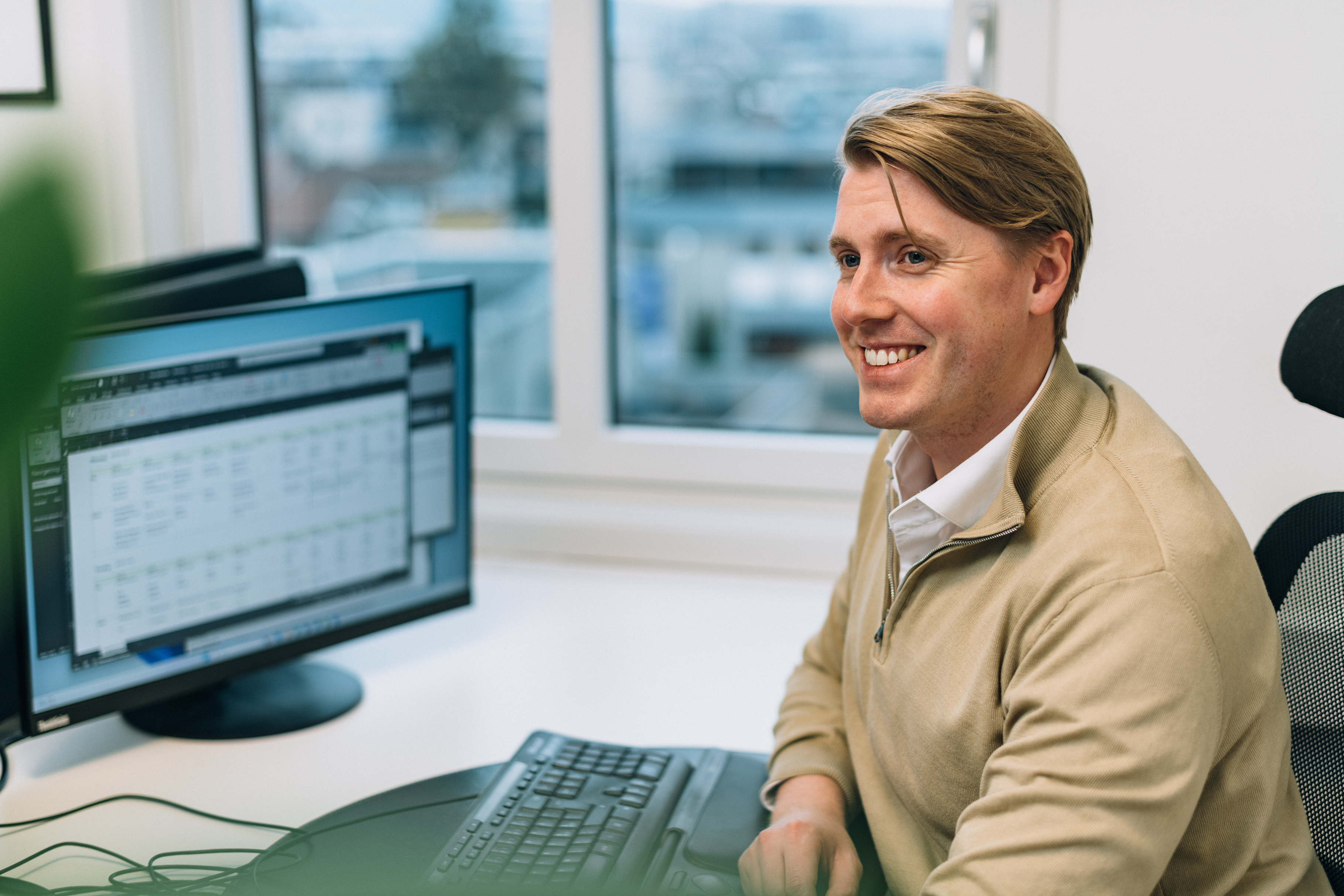 En person sitter ved et skrivebord foran en dataskjerm som viser et regneark eller en datatabell. Et vindu bak viser bygninger og trær utenfor.