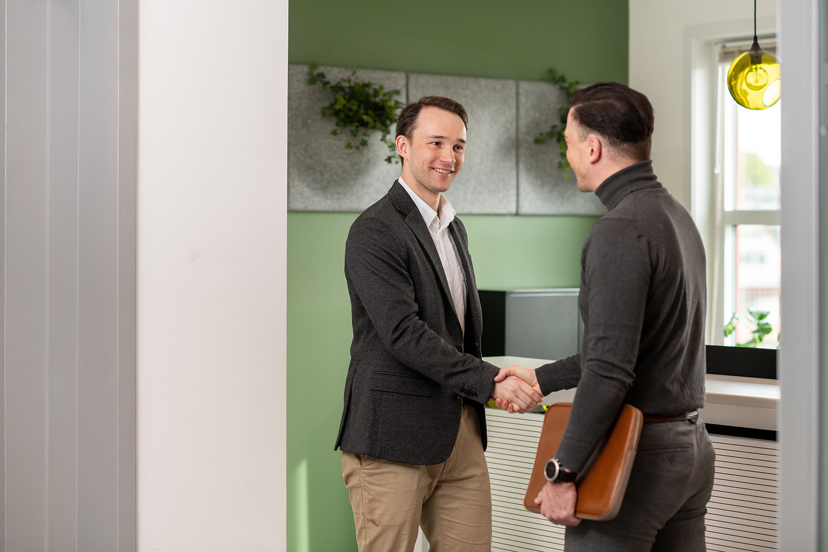 Two people shaking hands indoors, one holding a brown folder, with plants and a window in the background.