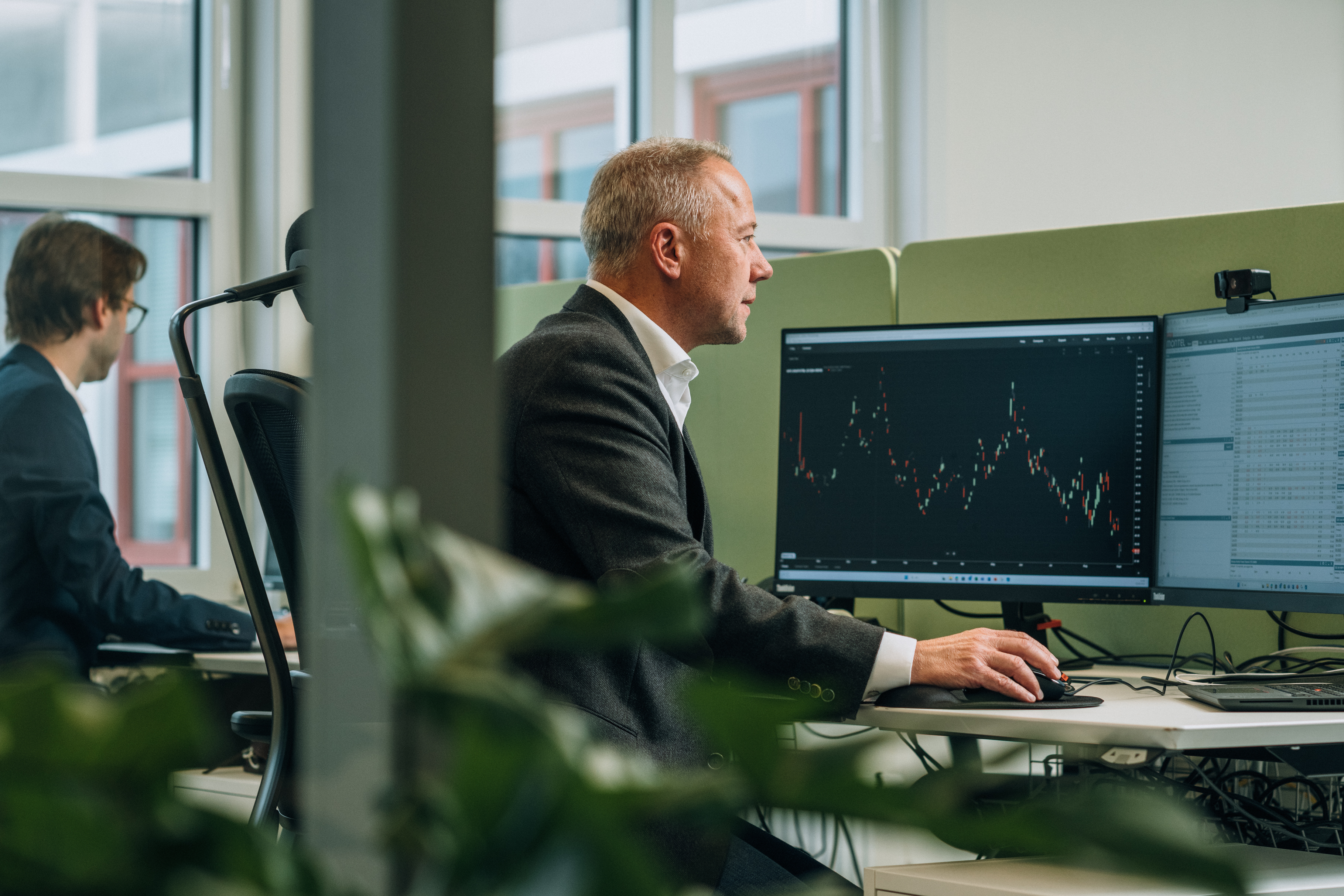 A man looks at a monitor displaying a financial chart with candlestick patterns in an office. Another man works at a desk in the background.