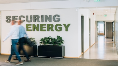 Two people walk through an office hallway with a "SECURING ENERGY" sign on the wall and potted plants below it.