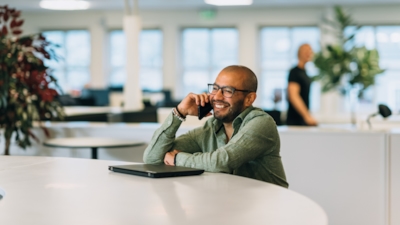 A man sits at a round table in a bright office, holding a phone to his ear. A closed laptop is on the table. A blurred person is walking by in the background.