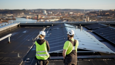 Two people in safety gear on a rooftop with solar panels, with a city and body of water in the background.