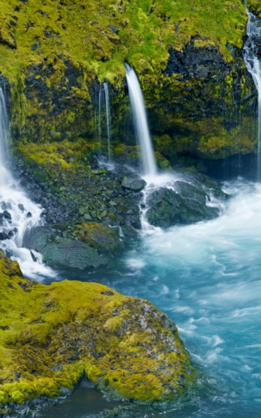 Small waterfalls cascade down a mossy cliff into a clear blue pool