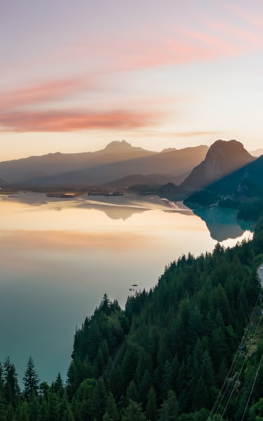Calm lake reflecting mountains and sky at sunrise or sunset, with a road along a forested slope.
